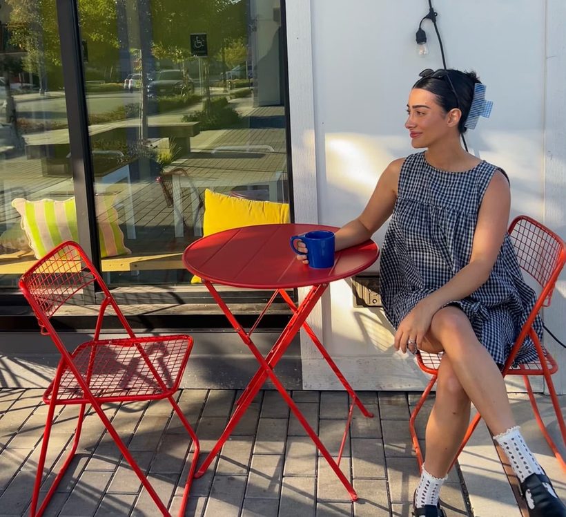 Bita at a small red cafe table in the sun, in profile, holding a blue mug