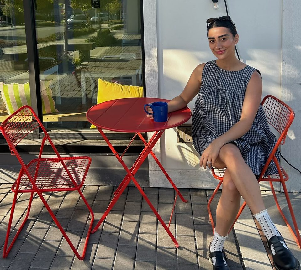 Bita at a small red cafe table in the sun, holding a blue mug, with an empty chair across from her