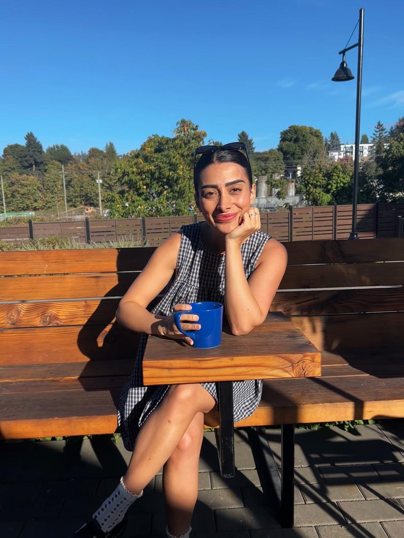 Bita sitting at an outdoor wooden table in the sun, smiling, with a blue mug